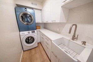A compact laundry area with a blue and white washing machine, a sink with a faucet, and a small basket. - Hillside Oasis with Jacuzzi, Ocean Views, BBQ, Treetop Master, EV Charger (San Diego)