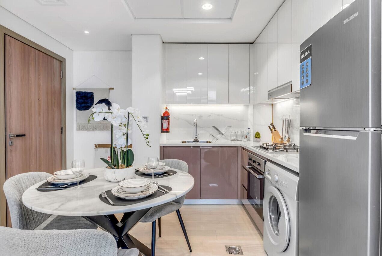 A modern kitchen with white cabinets, a stainless steel refrigerator, and a dining area with a round table and chairs.