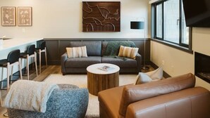 A modern living room with a sofa, coffee table, and ottoman, a dining area with chairs, and a TV mounted on the wall.