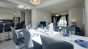 Een moderne eetruimte met een witte tafel voor vier personen, omringd door zwarte stoelen. Achtergrond toont een keuken met een koelkast en kasten.