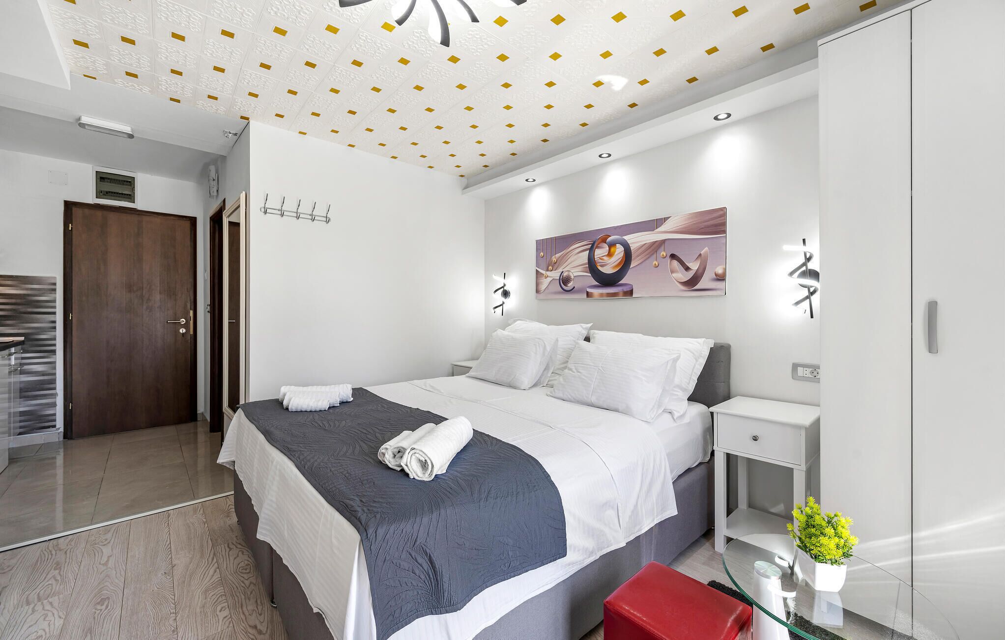Een moderne hotelkamer met een groot bed, een nachtkastje, een rode bank, een glazen tafel met een vaas en een plafond met decoratieve tegels.