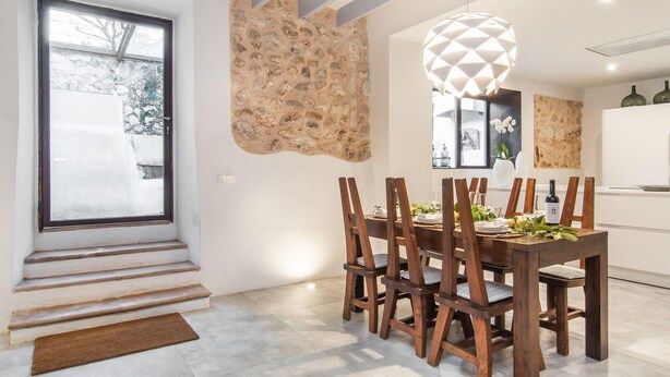 Comedor con mesa dispuesta para seis, sillas de madera, pared de acento de piedra, ventana grande y lámpara colgante.