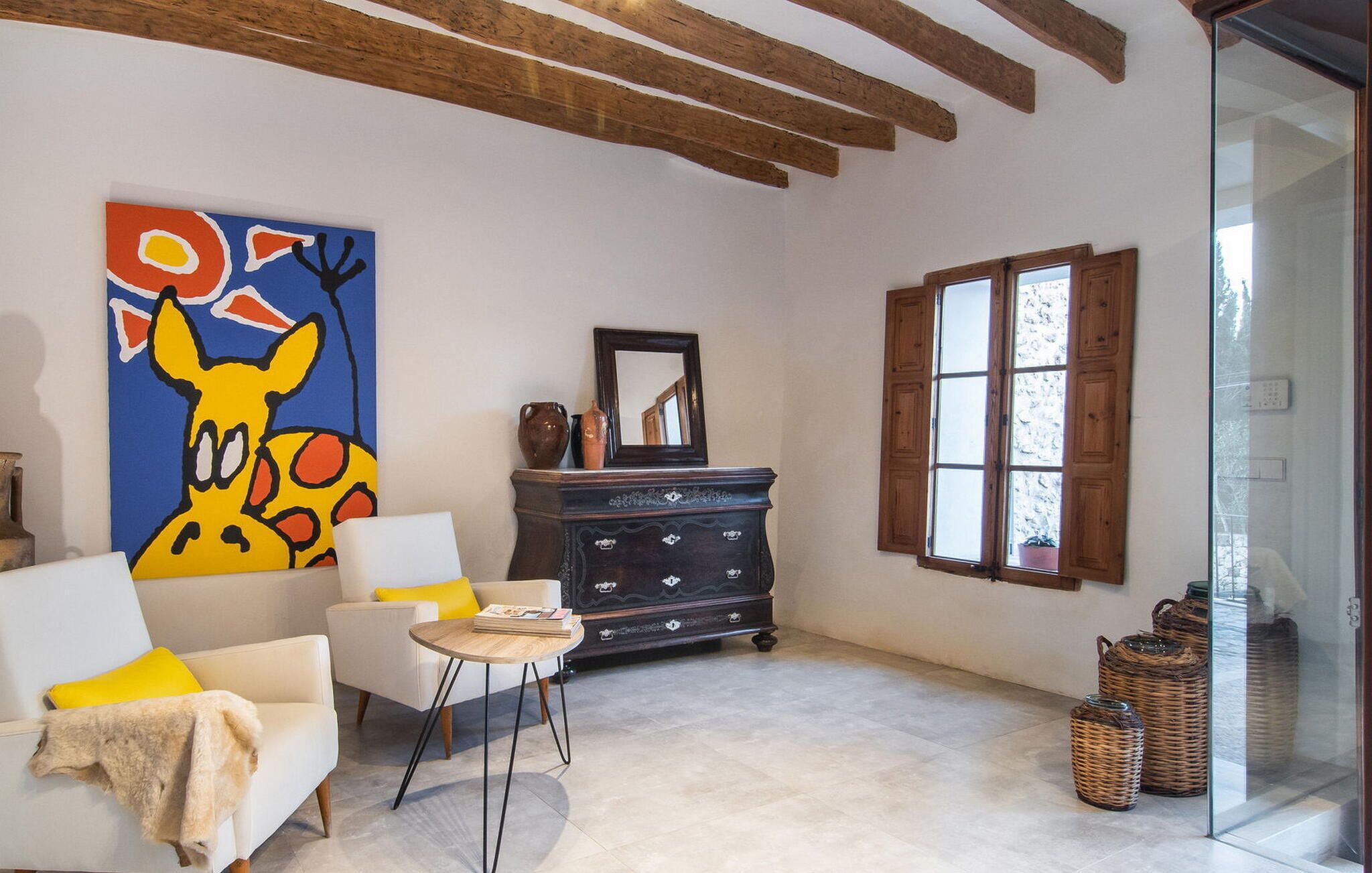 Uma sala de estar com sofá branco, uma mesinha redonda, um armário de madeira escura, um quadro abstrato colorido e uma janela com persianas de madeira.