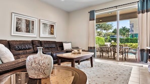 Ein modernes Wohnzimmer mit einer Ledersofa, einem Couchtisch und einer Vase. An der Wand befinden sich zwei gerahmte Bilder, und eine Glastür führt zu einem Außenbereich mit Pool und Gartenmöbeln.