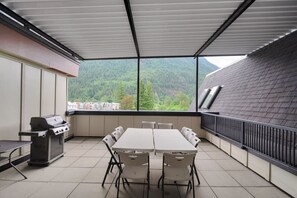 An outdoor patio with a table and chairs, a grill, and a view of a forested mountain.