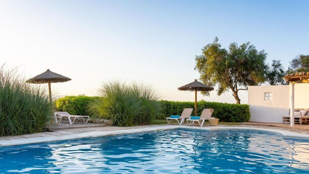 A swimming pool with lounge chairs and umbrellas, surrounded by greenery and a building. - Cozy apartment in Santa Margalida (Santa Margalida)