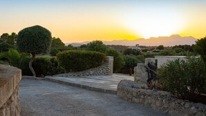 A paved pathway leading to a stone wall with a lamp post, surrounded by greenery and overlooking a mountainous landscape at sunset. - Cozy apartment in Santa Margalida (Santa Margalida)