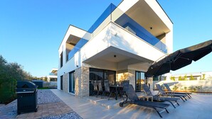 A modern two-story house with a flat roof, large windows, and a covered outdoor seating area with lounge chairs and a table.
