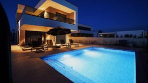 A modern two-story house with a swimming pool, outdoor seating, and a well-lit patio area.