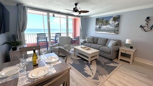 Een woonkamer met balkon en uitzicht op de oceaan, een eethoek met een tafel voor vier personen, en een grijze bank.