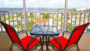 A balcony with a glass table and two red chairs, overlooking a marina and buildings. - Casa Playa #504 - Sea Horse (Fort Myers Beach)