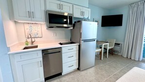 A modern kitchen with white cabinets, stainless steel appliances, a microwave, and a refrigerator. There is a dining area with a table and chairs, and a flat-screen TV mounted on the wall.