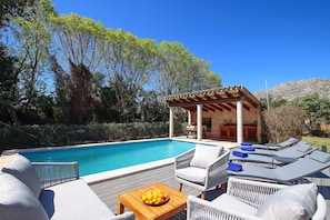 A poolside area with lounge chairs, a wooden table with fruit, and a pergola structure.