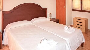A neatly made bed with white linens and a towel, a wooden headboard, a bedside table with a lamp, and a wooden dresser with drawers.