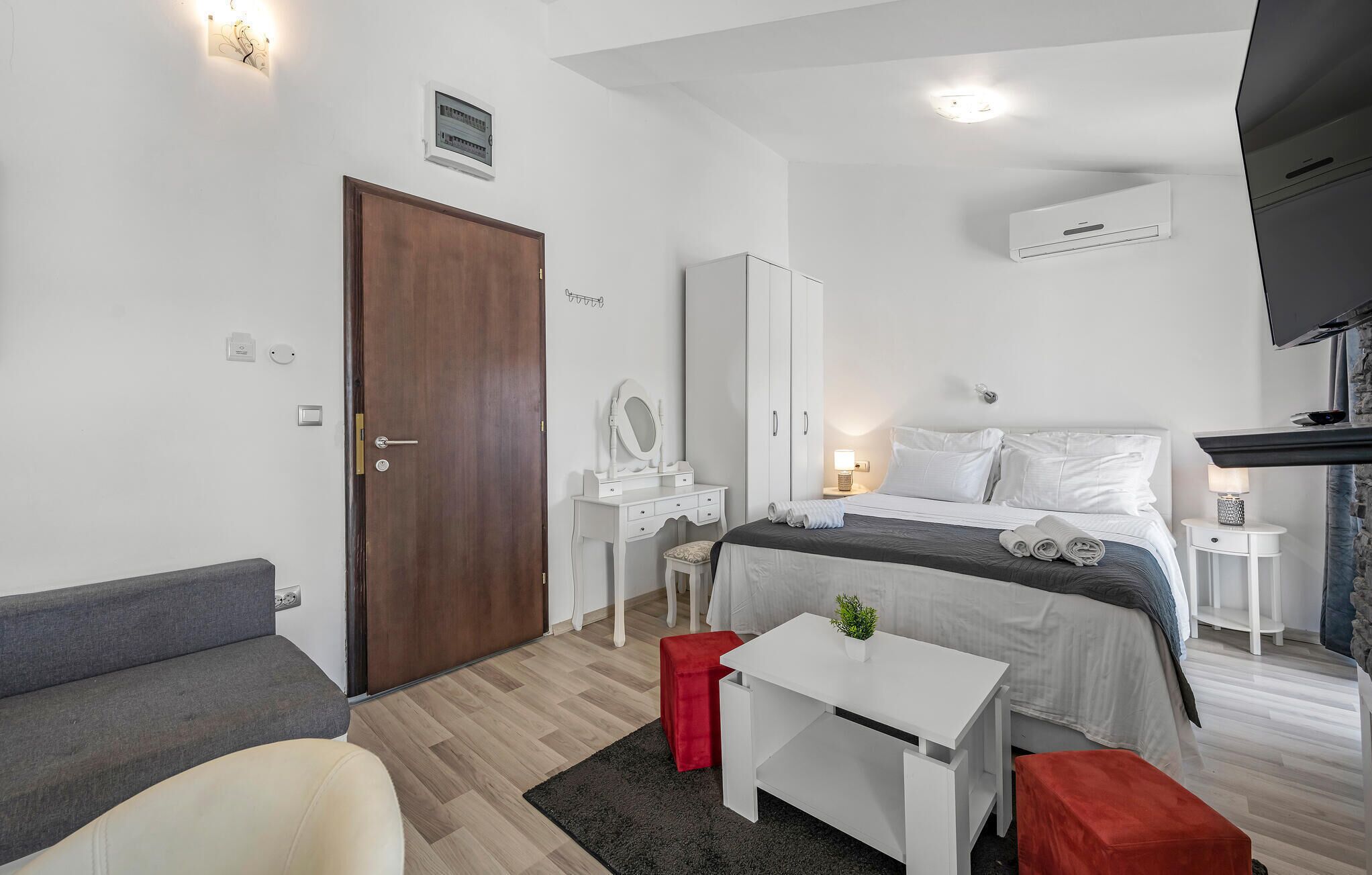 A modern hotel room with a wooden door, a white desk, a bed with white bedding, a red ottoman, and a flat-screen TV mounted on the wall.