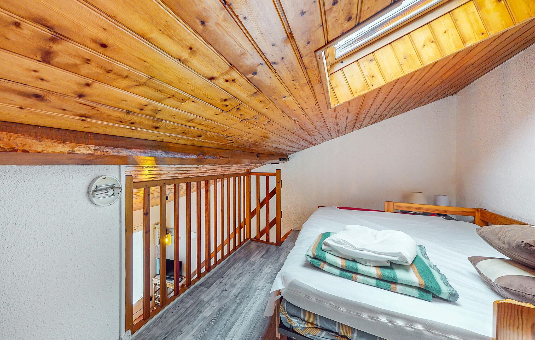 Une petite pièce chaleureuse avec un plafond en bois, un lit avec un matelas blanc et une couverture à rayures, un banc en bois et une petite table avec une lampe.
