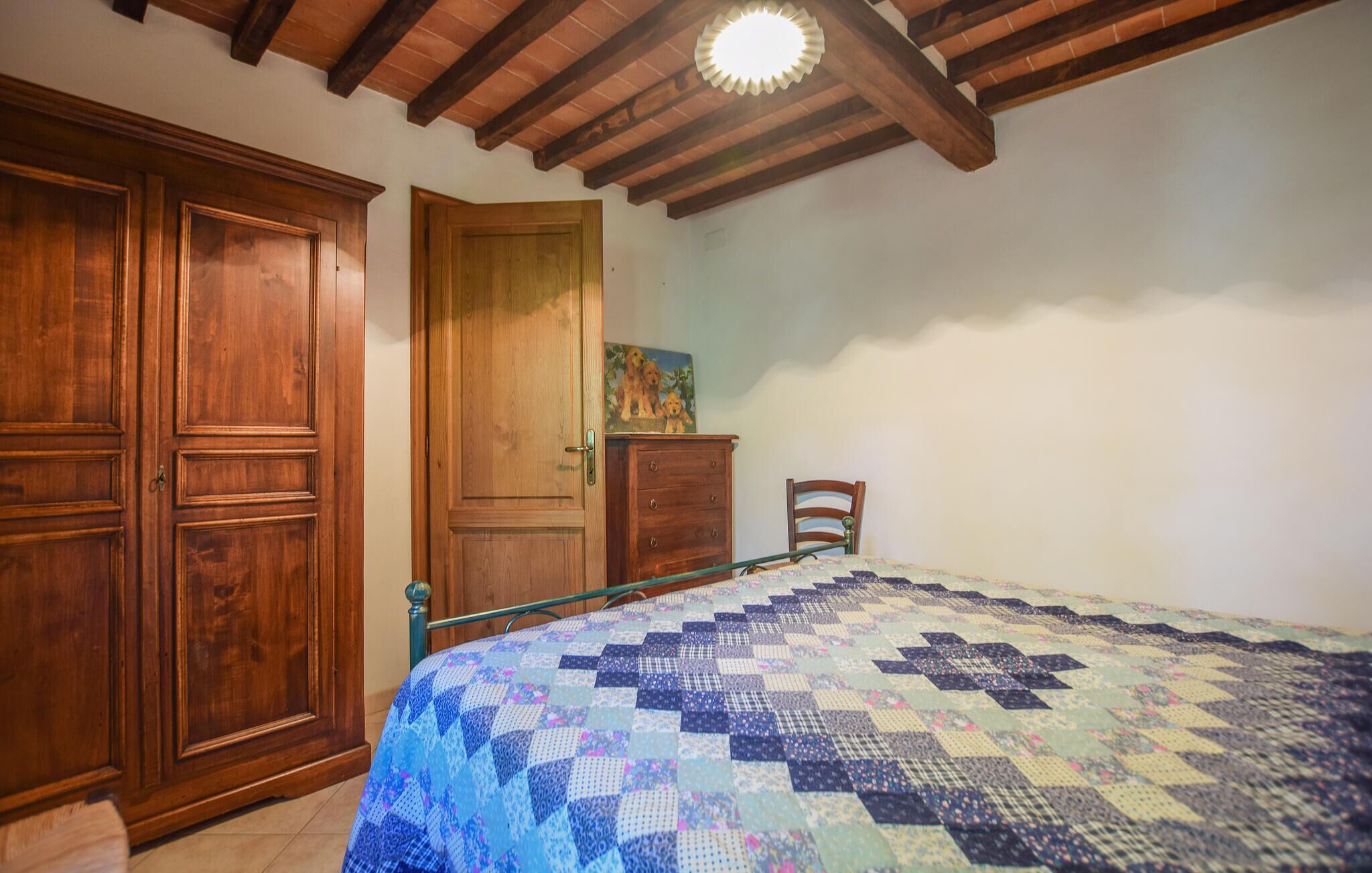 A bedroom with a wooden wardrobe, a bed with a quilt, a wooden dresser, and a painting on the wall.