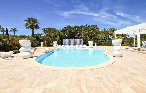 A swimming pool with white lounge chairs and decorative planters, surrounded by palm trees and a clear sky.