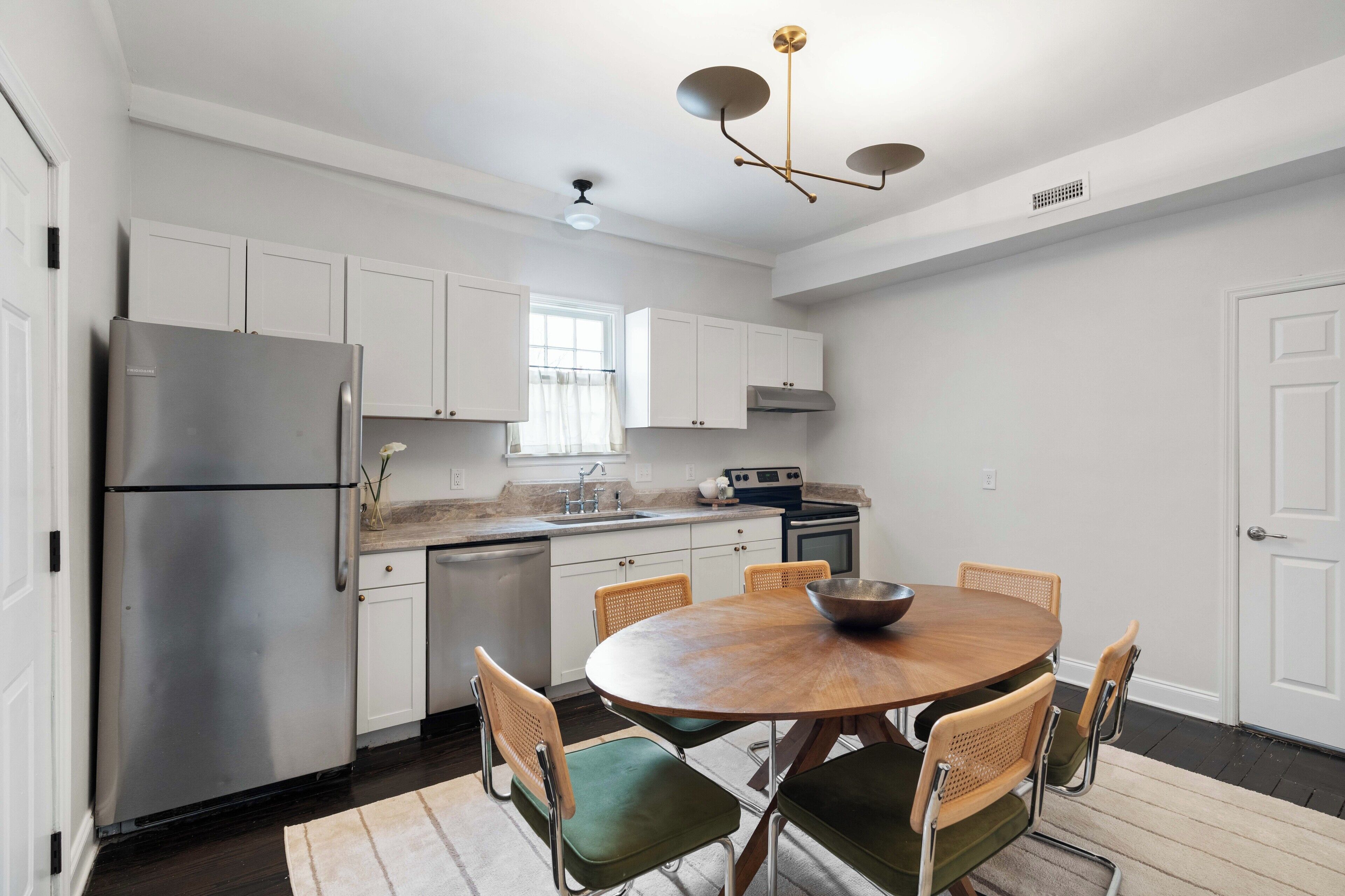 A modern kitchen with white cabinets, a stainless steel refrigerator, a dining table with chairs, and a pendant light.