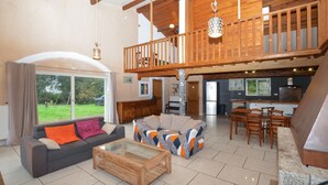 A spacious living area with a sofa, a coffee table, and a dining set. A balcony with wooden railings and hanging lights is visible.