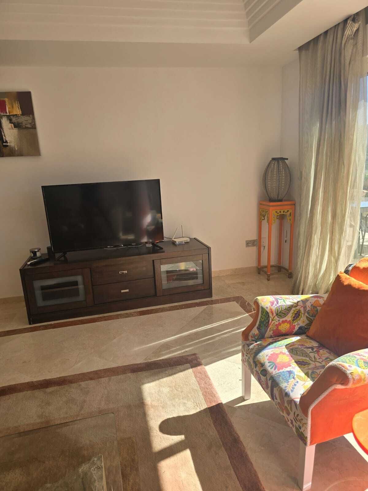 Ein Wohnzimmer mit einem Flachbildfernseher auf einem dunklen Holzschrank, einem farbenfrohen Sofa, einem kleinen Beistelltisch mit einer Vase und einem großen Fenster mit transparenten Vorhängen.