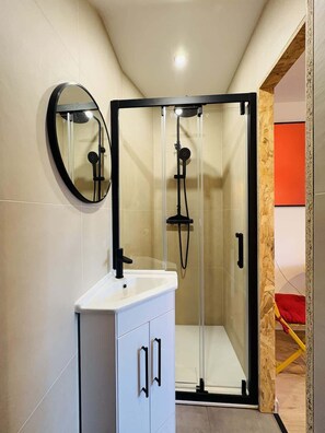 Ein modernes Badezimmer mit Duschkabine aus Glas, einem weißen Waschbecken mit schwarzen Griffen und einem runden Spiegel.