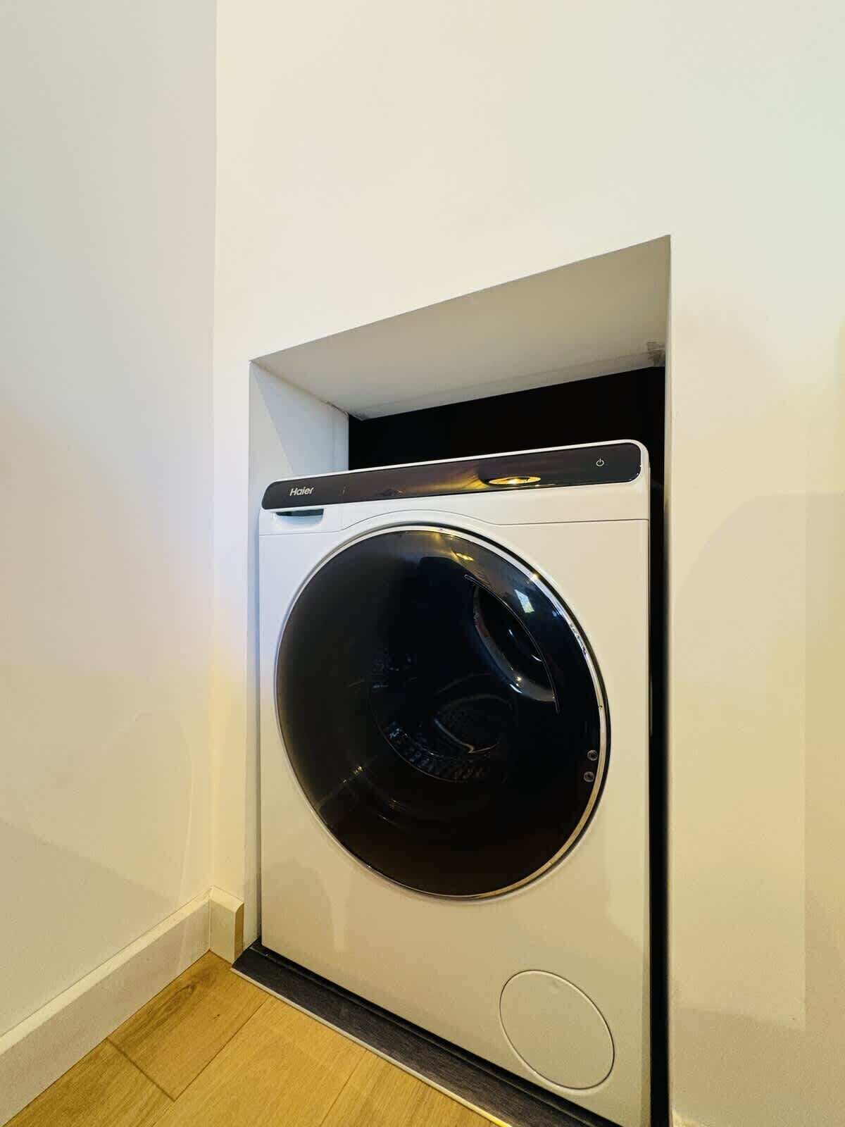 A compact, wall-mounted washing machine with a control panel on top, situated in a corner of a room with wooden flooring.