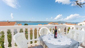 Un balcón con sillas y una mesa blancos, con vistas a la costa, edificios y un cielo despejado.