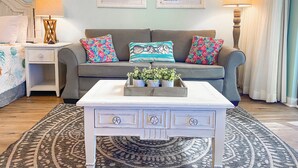Una sala de estar con un sofá gris, almohadas coloridas, una mesa de centro blanca con una planta y dos obras de arte enmarcadas sobre el sofá.