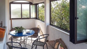 En lille terrasse med et rundt bord og stole, en potteplante og udsigt til træer gennem store vinduer.