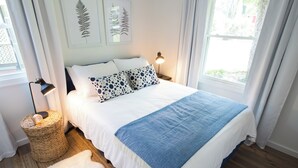 Una cama bien hecha con una manta blanca y azul, un cojín estampado y una pequeña lámpara de noche. Hay una cesta tejida en el suelo al lado de la cama.