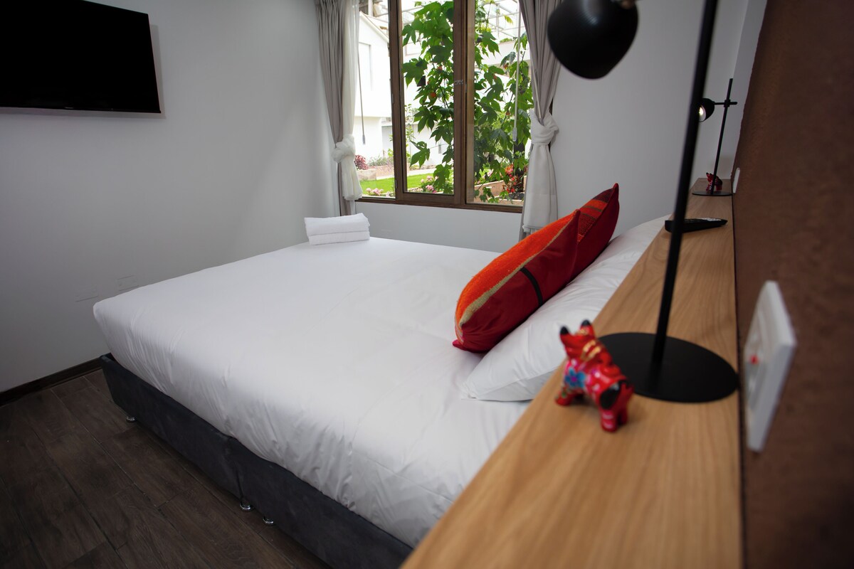 Una cama bien hecha con ropa de cama blanca y un cojín rojo, una figurita de juguete en la mesita de noche y una ventana con cortinas.