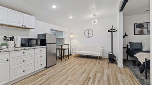 Cocina moderna con armarios blancos, microondas y refrigerador. En la sala de estar, hay un sofá blanco y una cama con estructura negra.