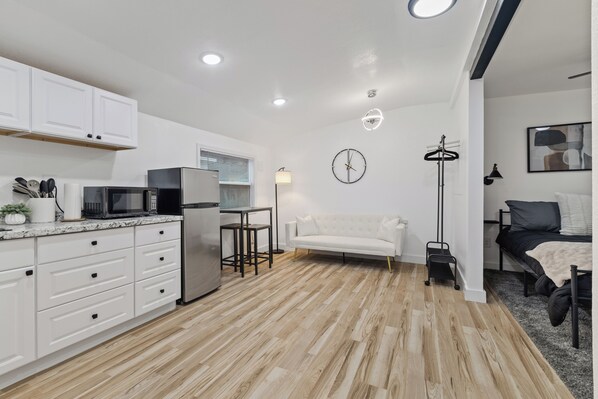 Cocina moderna con armarios blancos, microondas y refrigerador. En la sala de estar, hay un sofá blanco y una cama con estructura negra.