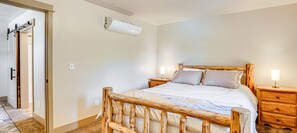Une chambre à coucher avec un cadre de lit en bois, deux tables de chevet avec des lampes, un climatiseur fixé au mur et une porte donnant sur une autre pièce.
