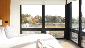 Une chambre moderne avec un grand lit, une poutre en bois au plafond et de grandes fenêtres donnant sur un paysage arboré et un plan d’eau.