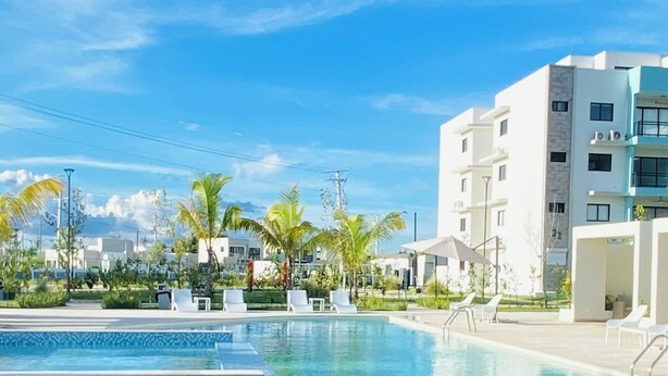A swimming pool with lounge chairs, palm trees, and modern buildings in the background. - Stylish and Modern Apartment in Punta Cana (Punta Cana)