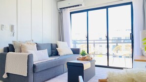 A modern living room with a grey sofa, a small side table, and a view of the city through large windows. - Stylish and Modern Apartment in Punta Cana (Punta Cana)