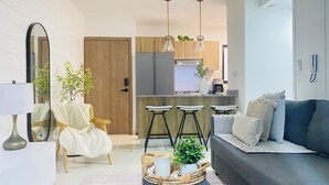 A modern living room with a sofa, a chair, a coffee table with a plant, and a kitchen area with bar stools and a refrigerator. - Stylish and Modern Apartment in Punta Cana (Punta Cana)