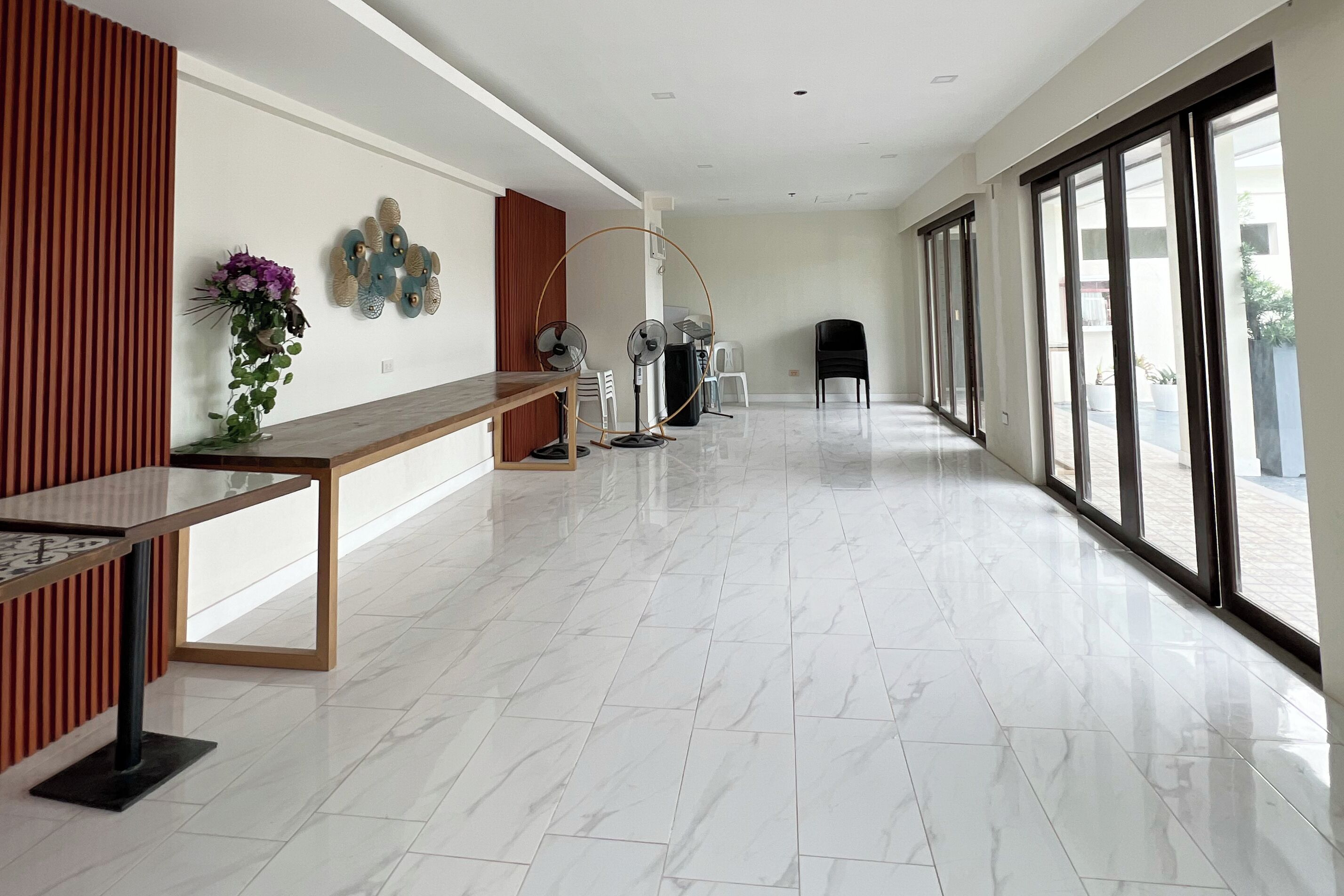 A spacious hallway with a reception desk, a wooden panel wall, a vase with flowers, two wall-mounted fans, and a large window with a view of the outside.