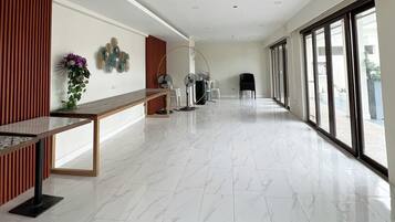 Un hall espacioso con mostrador de recepción, un revestimiento de madera, un jarrón con flores, dos ventiladores de pared y un amplio ventanal con vistas al exterior.
