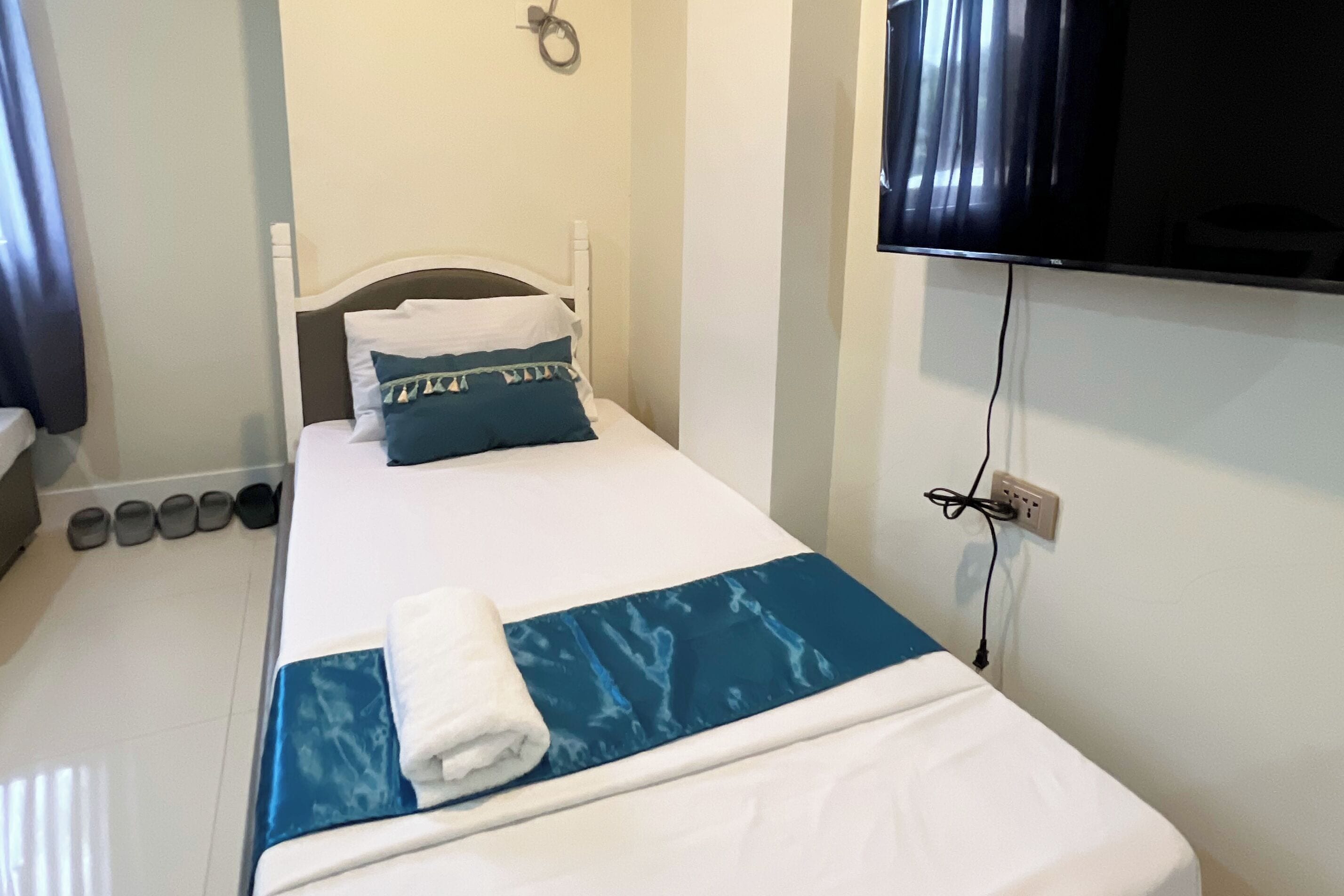 A neatly made bed with a blue and white bedspread, a white pillow, and a folded white towel. There is a television mounted on the wall to the right of the bed.