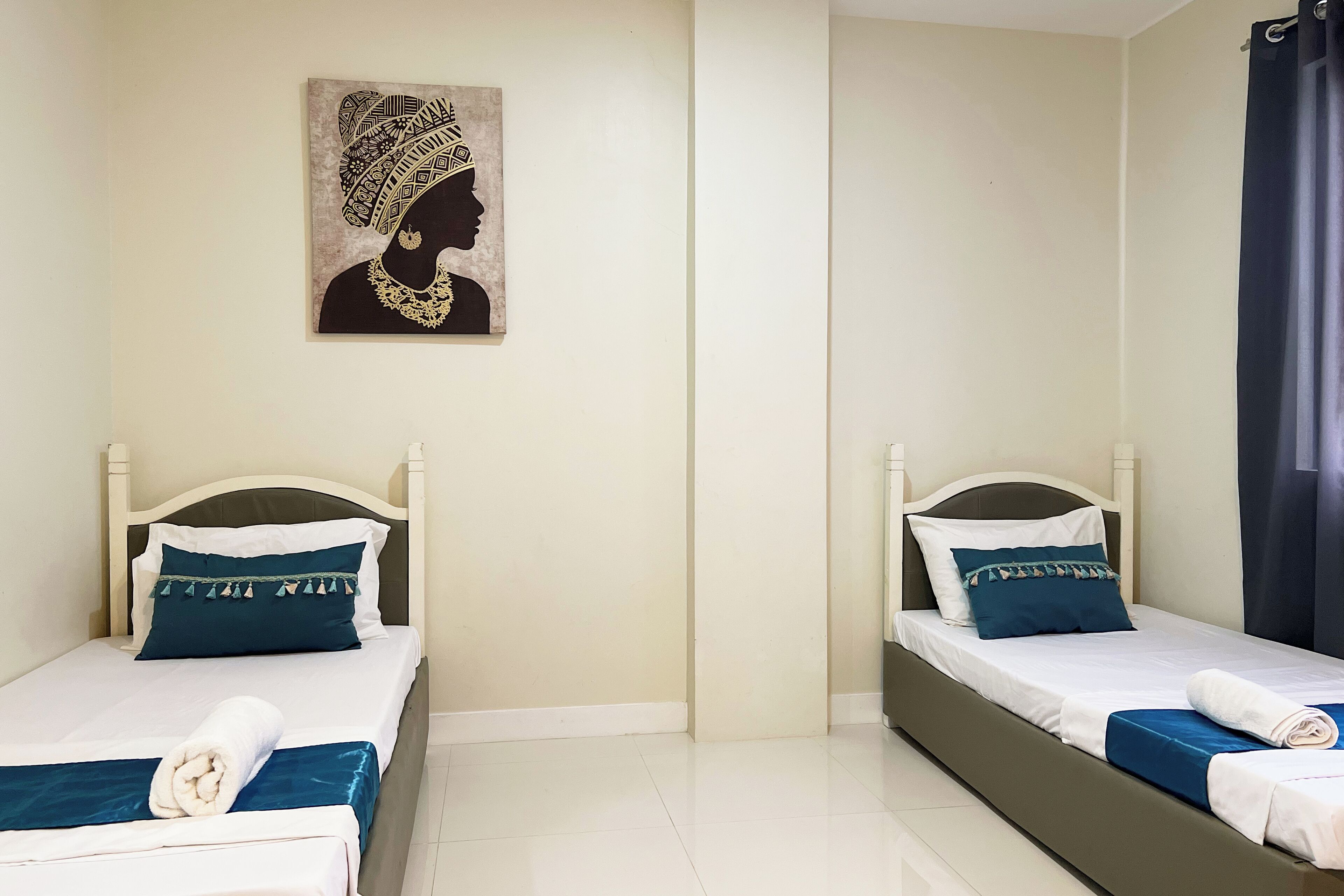 Two twin beds with white headboards, blue bedding, and a rolled towel on each bed. A framed artwork is on the wall above the beds.