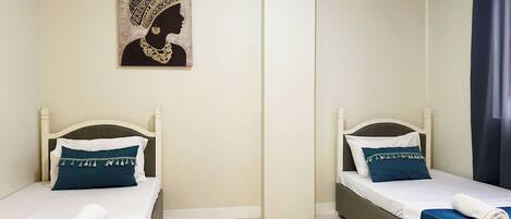 Two twin beds with white headboards, blue bedding, and a rolled towel on each bed. A framed artwork is on the wall above the beds.