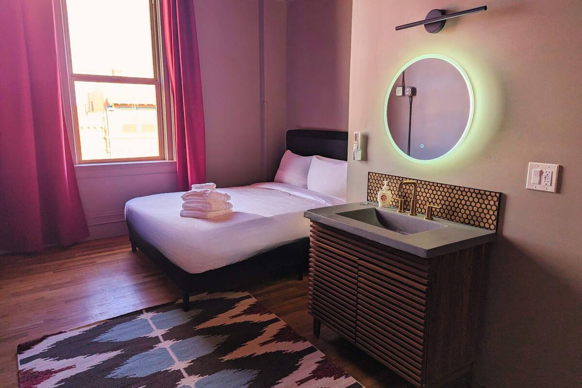 Habitación de hotel con cama, mesita de noche con espejo, lavabo y ventana con cortinas.