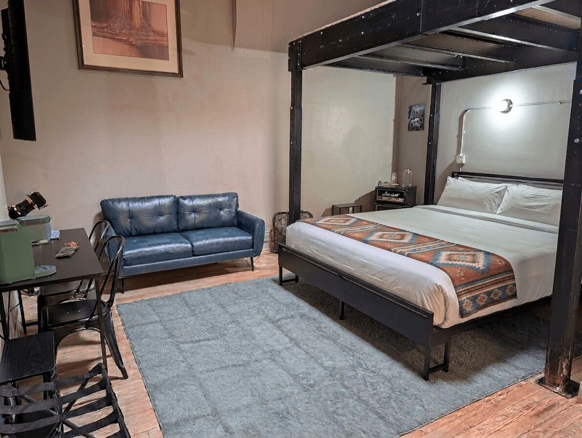 A bedroom with a four-poster bed, a blue sofa, a desk with chairs, and a small table with a lamp.