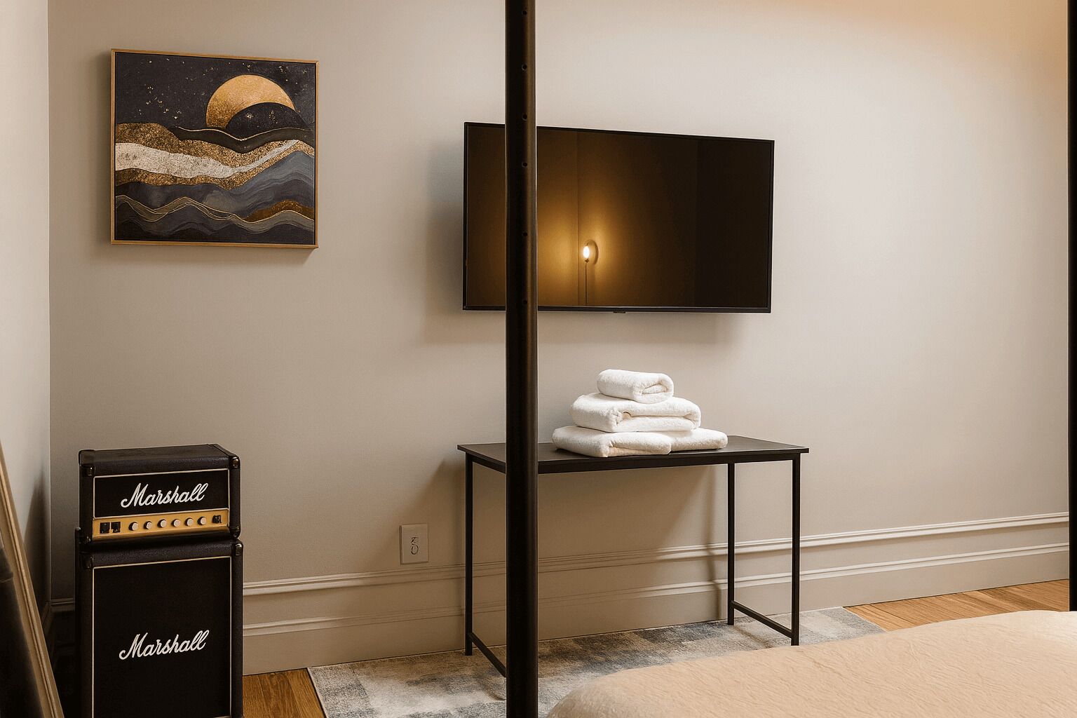 A hotel room with a flat-screen TV mounted on the wall, a small table with folded towels, a Marshall amplifier, and a painting of a moonlit sea.