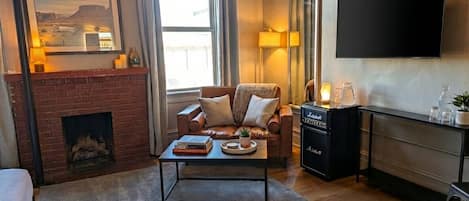 A cozy living room with a fireplace, a comfortable sofa, a coffee table with books and a plant, a TV mounted on the wall, and a console table with a speaker.