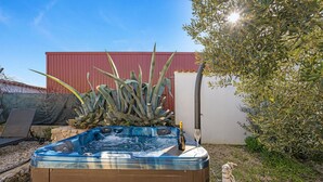 Un bain à remous avec une bouteille de champagne et un verre, entouré de plantes et avec un bâtiment rouge en arrière-plan.