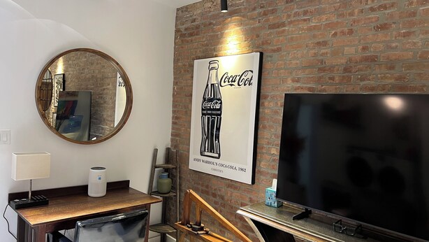 A modern living room with a brick wall, a large flat-screen TV, a round mirror, a wooden desk, and a coffee table.